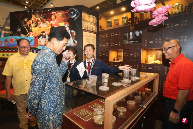 在南桥中学学生导览员的提议下，教育部长王乙康（左二）在有互动元素的“落地生根，开拓新家园”展览中医展区，举起装有中药材的塑料杯子嗅了嗅药香，左一为宗乡会馆联合总会会长陈奕福。（梁伟康摄）