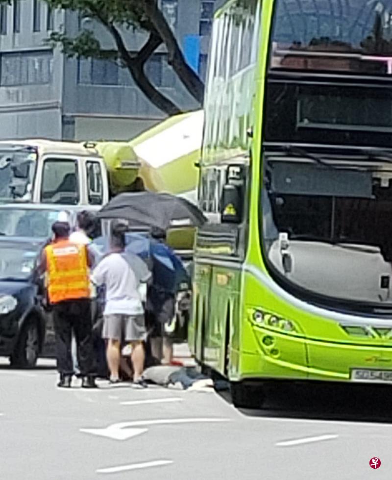 伤者倒在巴士右侧后方，好心父子帮忙撑伞。（受访者提供）